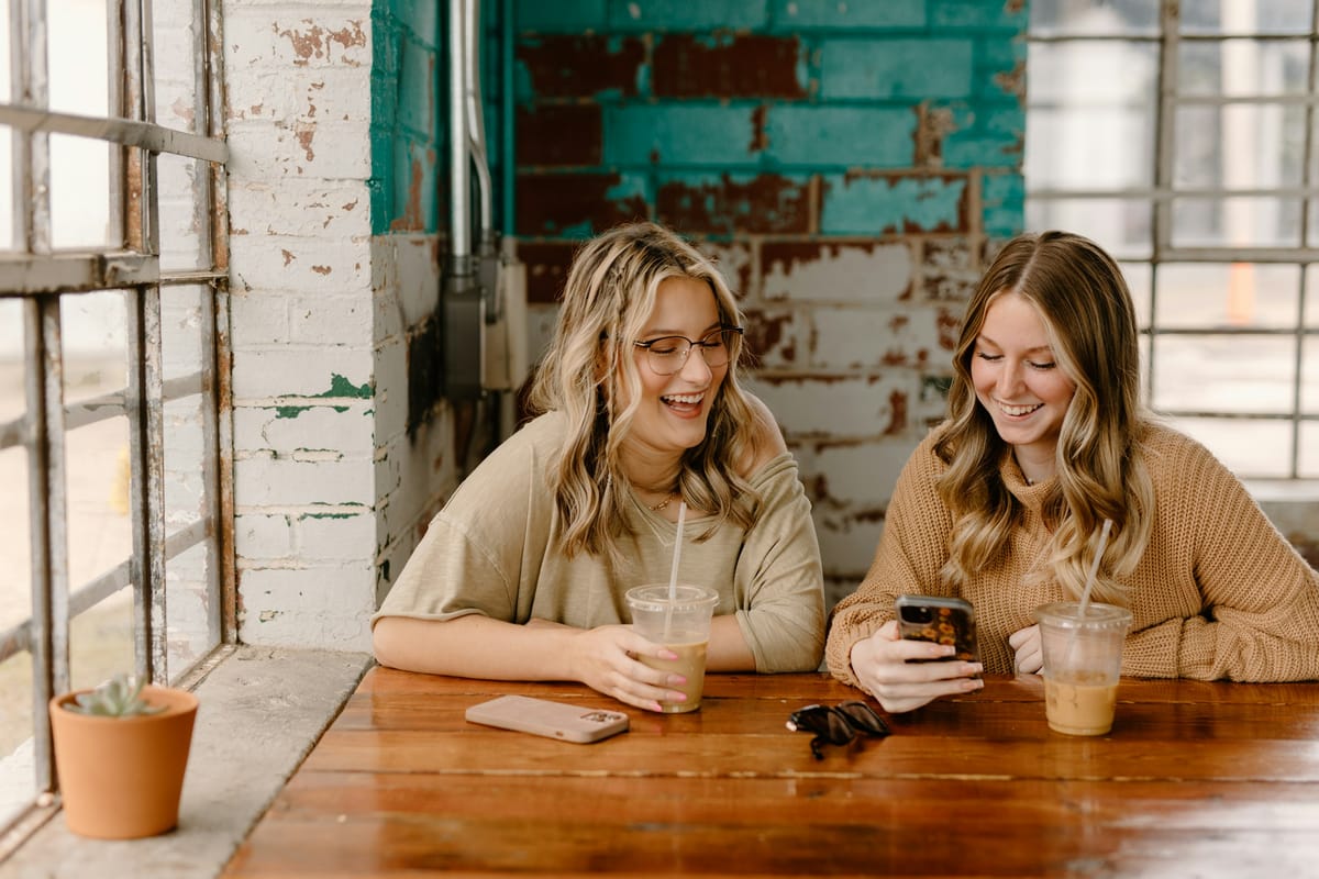 Two friends laughing together over coffee while one shows the other her phone